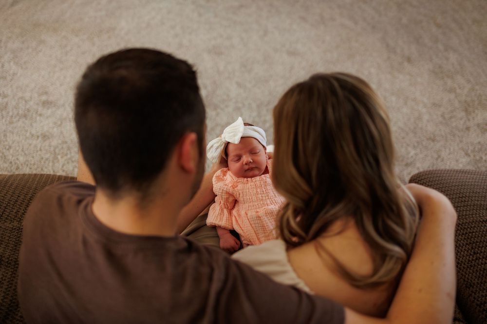 Parents holding their newborn baby girl in a pink knit wrap and white bow headband on a cozy sofa.