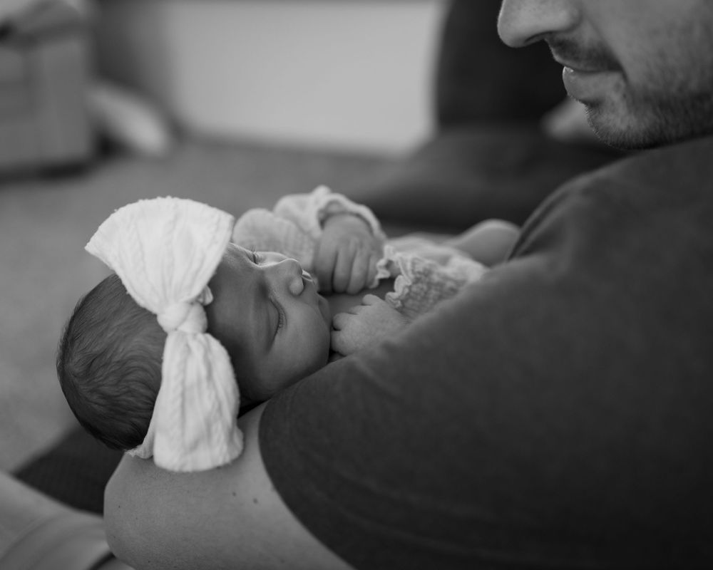 Black and white photo of a father cradling his newborn baby wearing a white knotted hospital hat.
