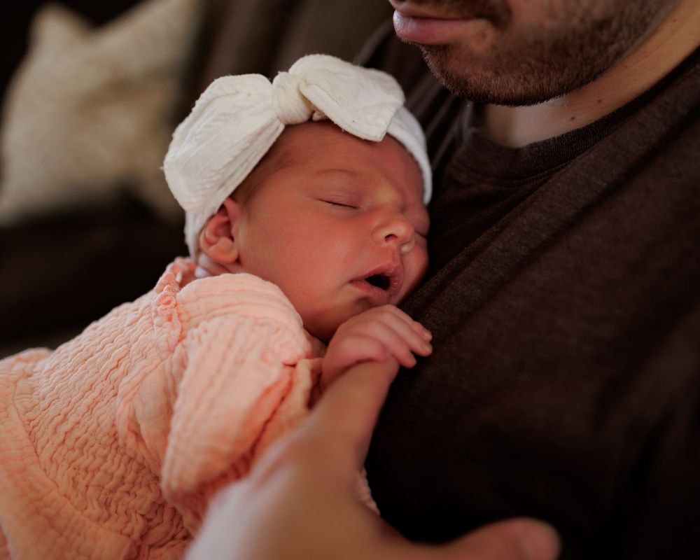 Newborn baby girl in pink outfit and white bow headband sleeping peacefully in fathers arms.