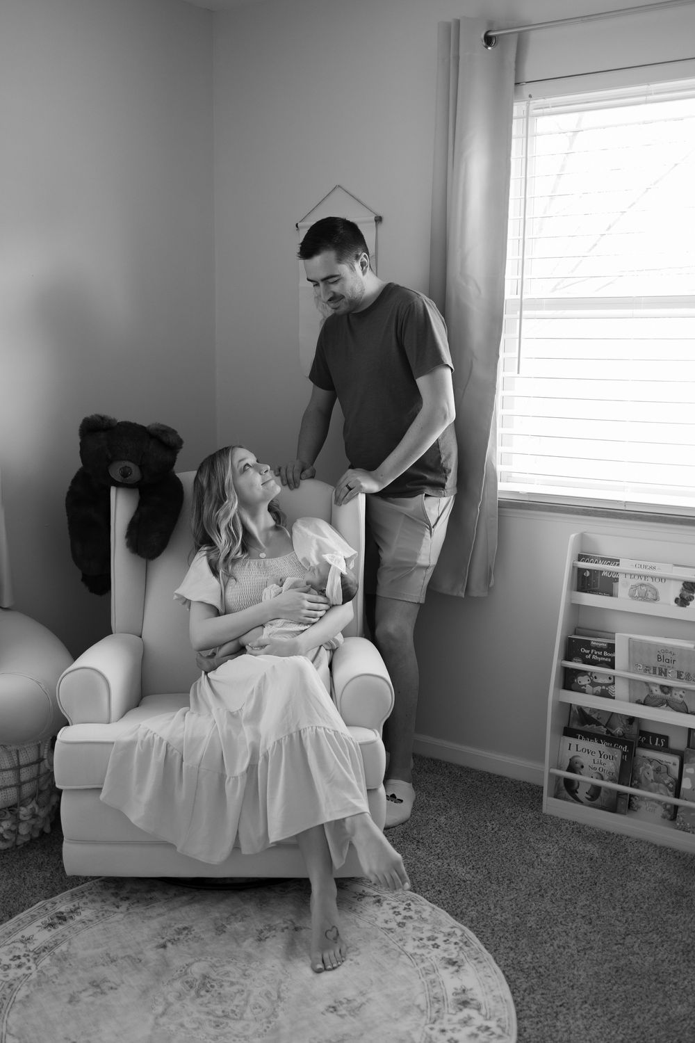 Black and white photo of a couple in a nursery, woman sitting in glider holding newborn baby while man stands beside her.