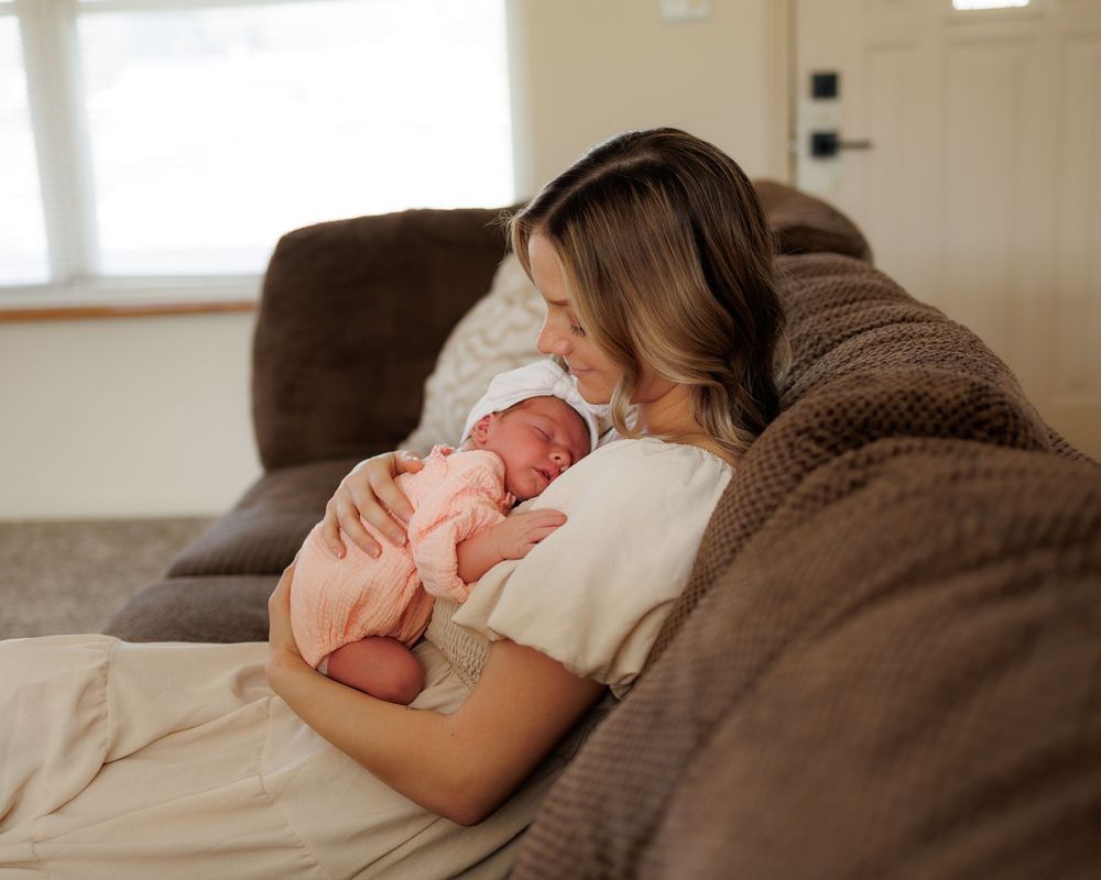 A mother tenderly holds her newborn baby while resting on a brown sofa in a cozy home setting.