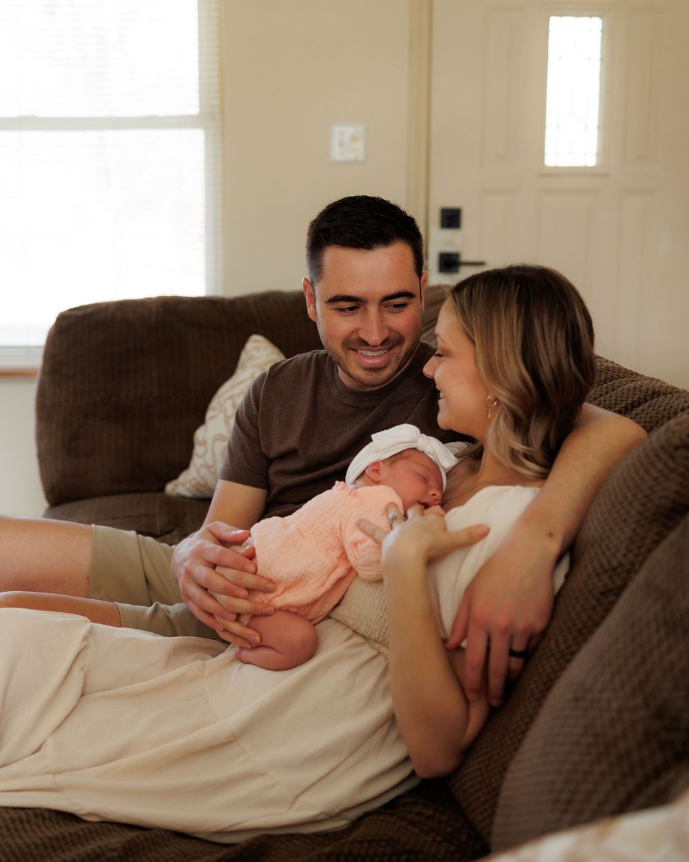 A couple smiles lovingly at their newborn baby while sitting together on a cozy brown sofa at home.