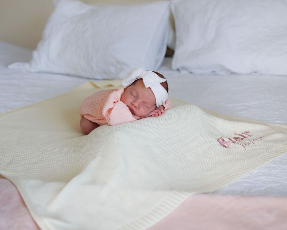 Newborn baby sleeping peacefully on a cream blanket with a white bow headband on a bed.
