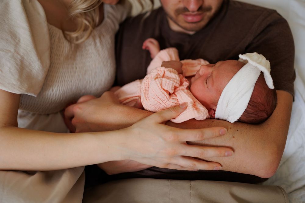 Parents cradling their newborn baby wrapped in a pink knit blanket and white bow headband.