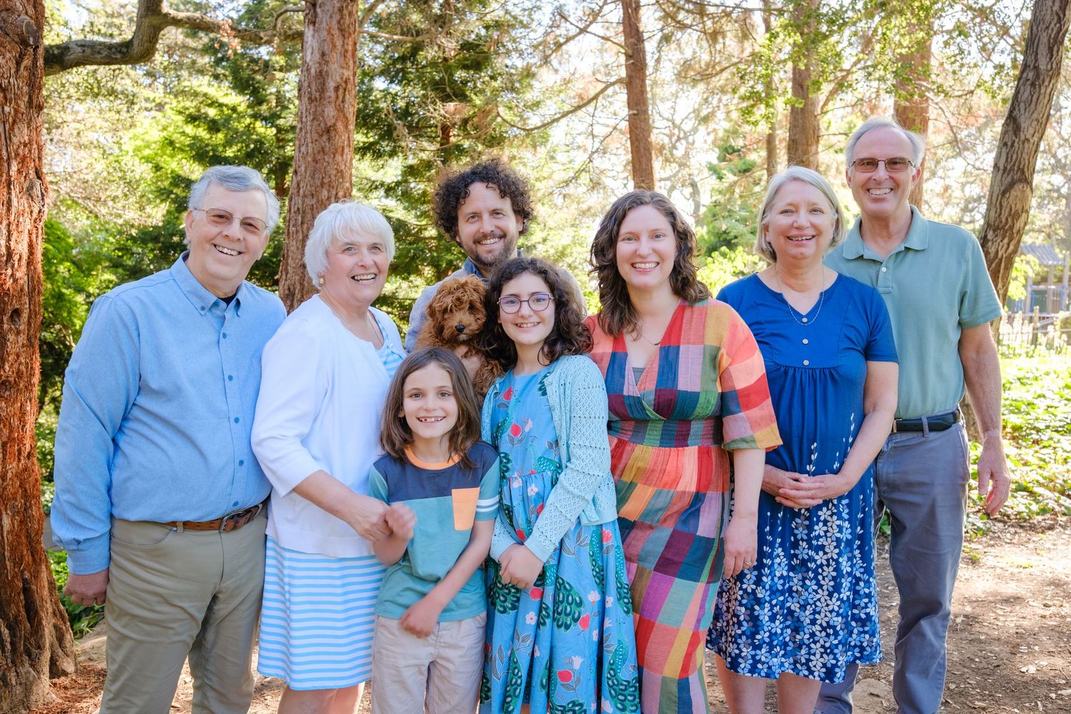 Multi-generational family group smiling outdoors in a sunny garden setting with tall trees and a small dog.