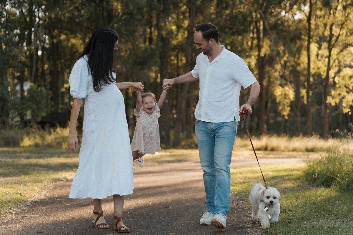 Happy family with toddler and white dog enjoy a sunny walk on a park path surrounded by trees in rouse hill regional park maternity session