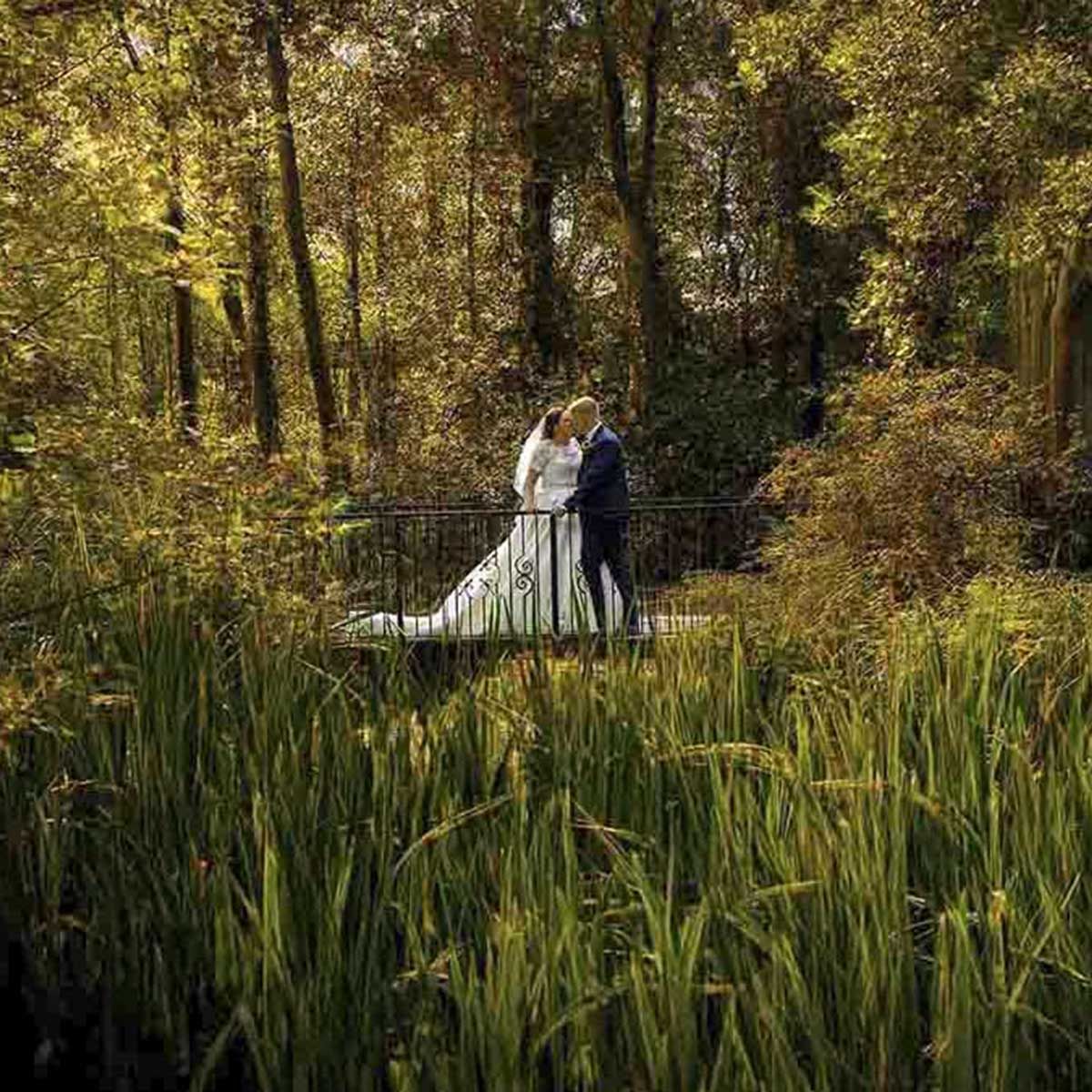 A bride and groom share a romantic kiss on a bridge surrounded by lush green trees and tall grass in a garden setting.