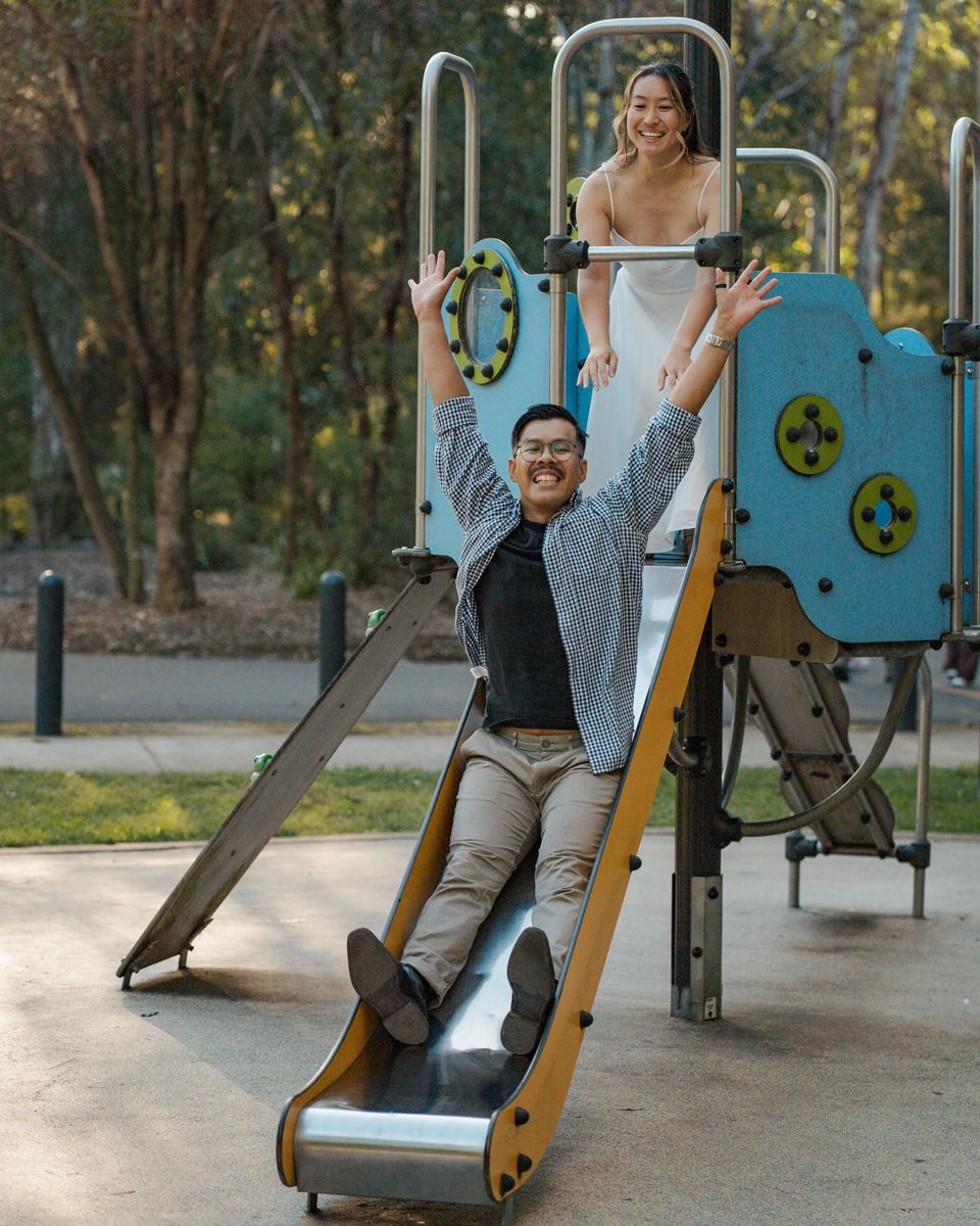 Couple playing on a colorful playground slide in a park, man sliding down with arms raised, woman smiling above couples photoshoot in sydney