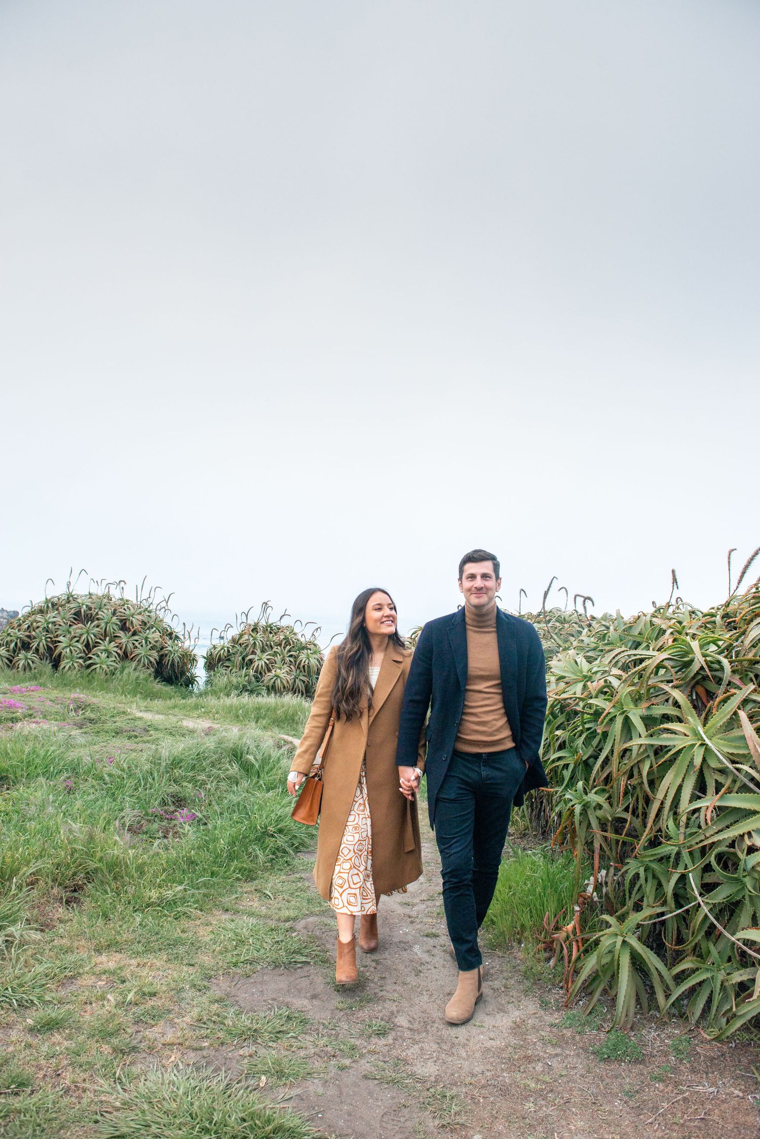 Couple holding hands walking on a scenic coastal path, woman in camel coat and man in dark teal sweater and navy pants.