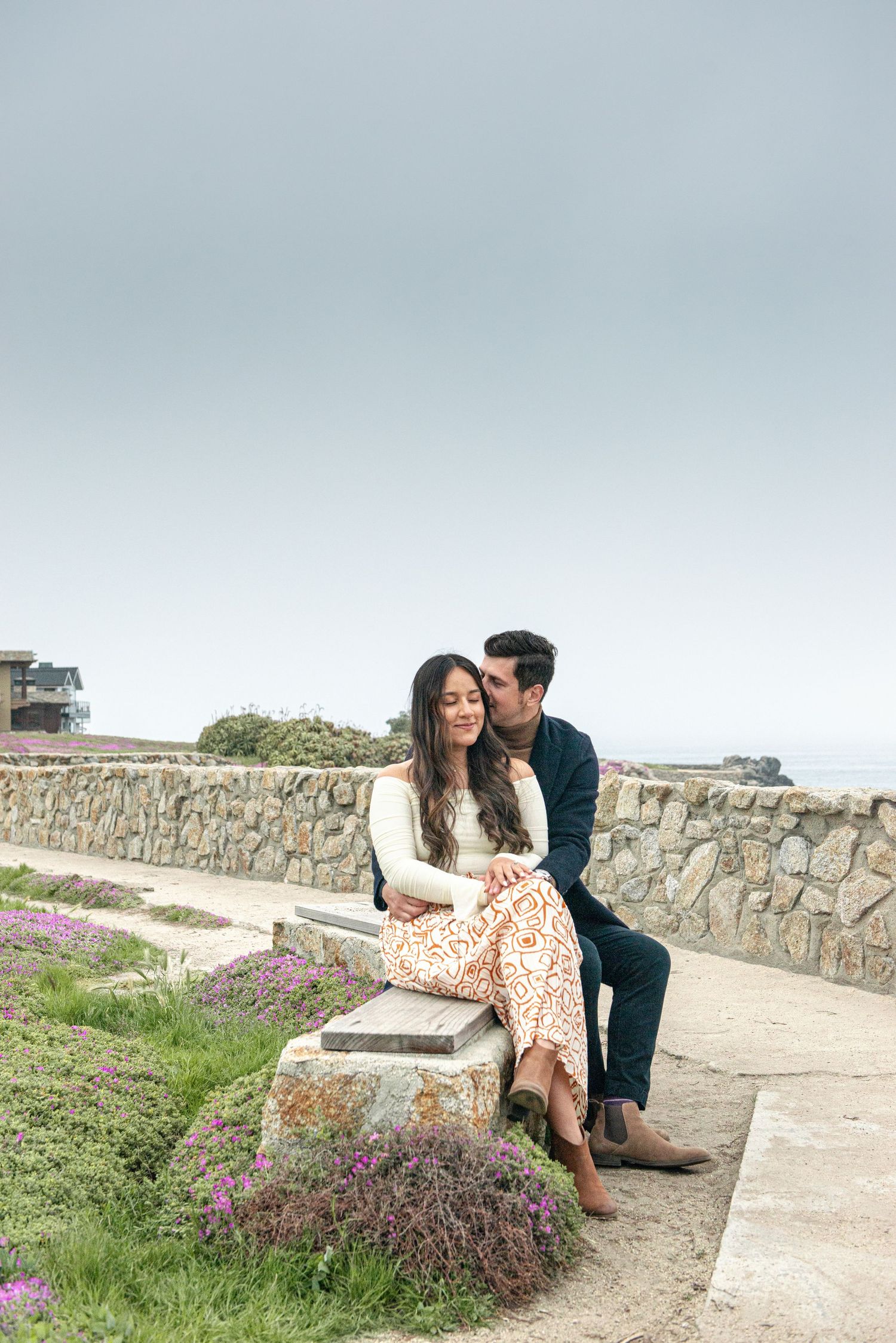 Couple sitting on a stone bench near a coastal wall with purple wildflowers, sharing a tender moment.