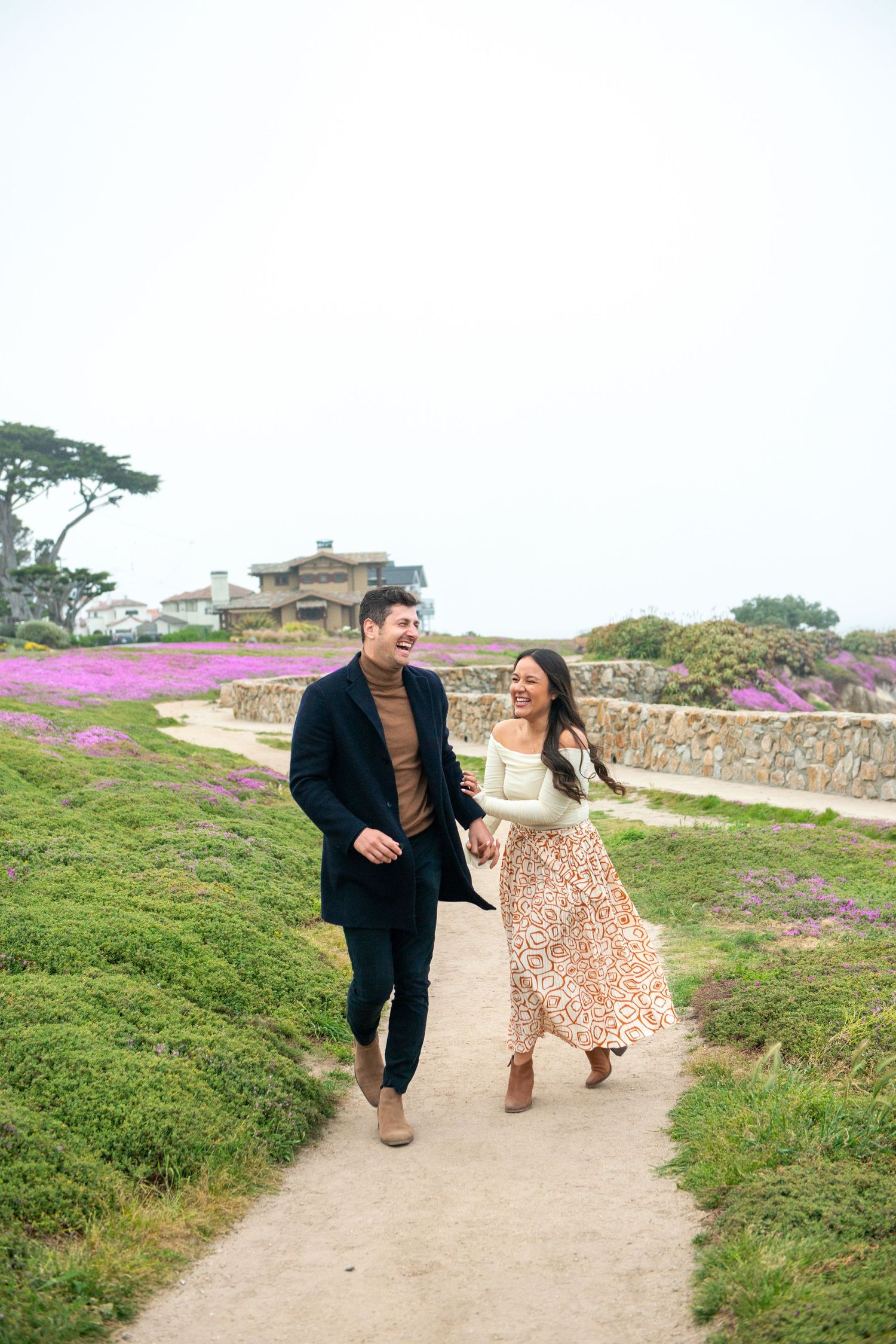 A couple walks hand-in-hand on a coastal path surrounded by purple wildflowers and a stone wall on a misty day.