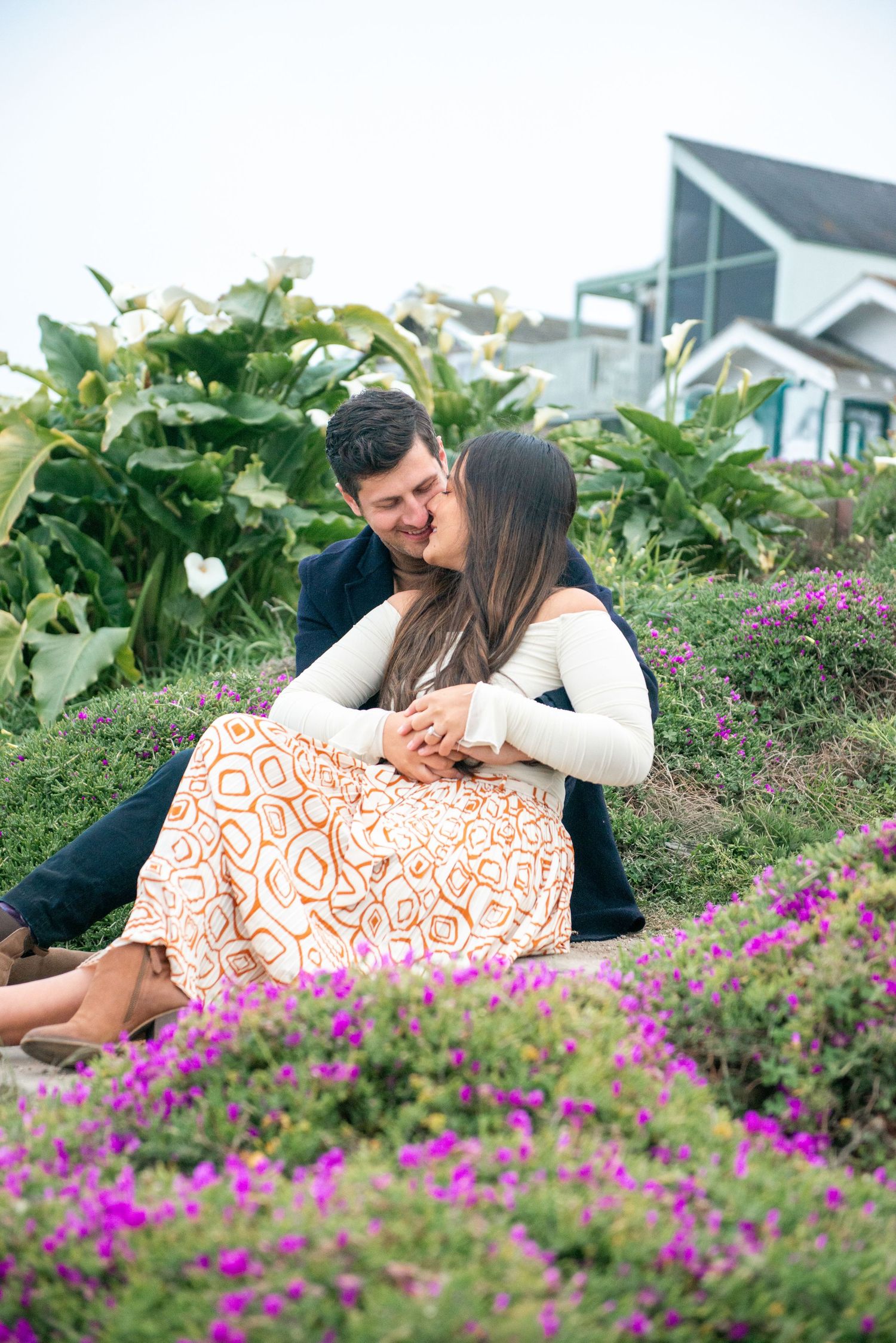 Couple sharing a kiss while sitting among purple and green flowers with a coastal house in the background.