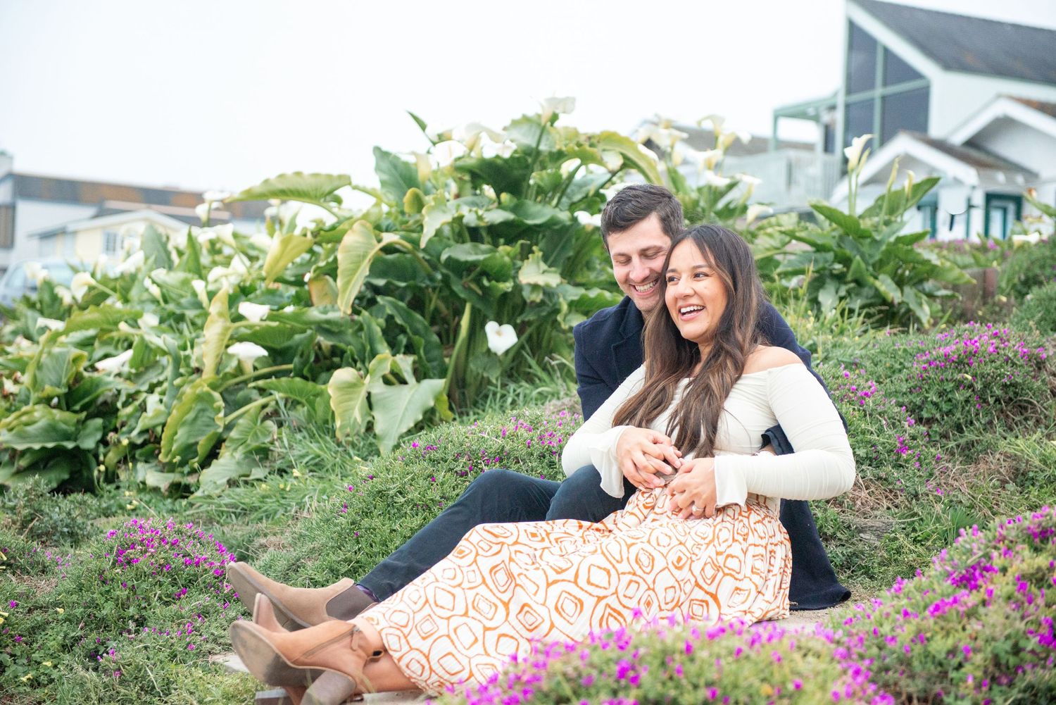 Happy couple sitting together on a hillside garden with lush green plants and purple flowers in background.