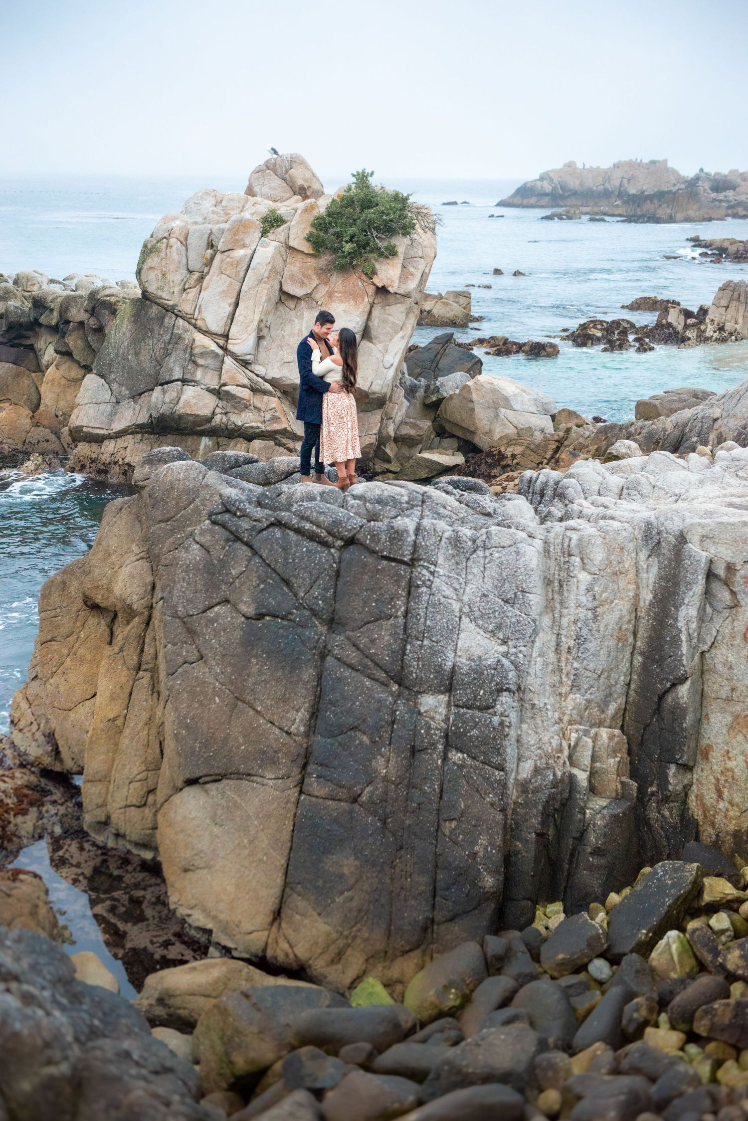 Couple embracing on rocky coastal cliffs with ocean waves and rock formations in the background.