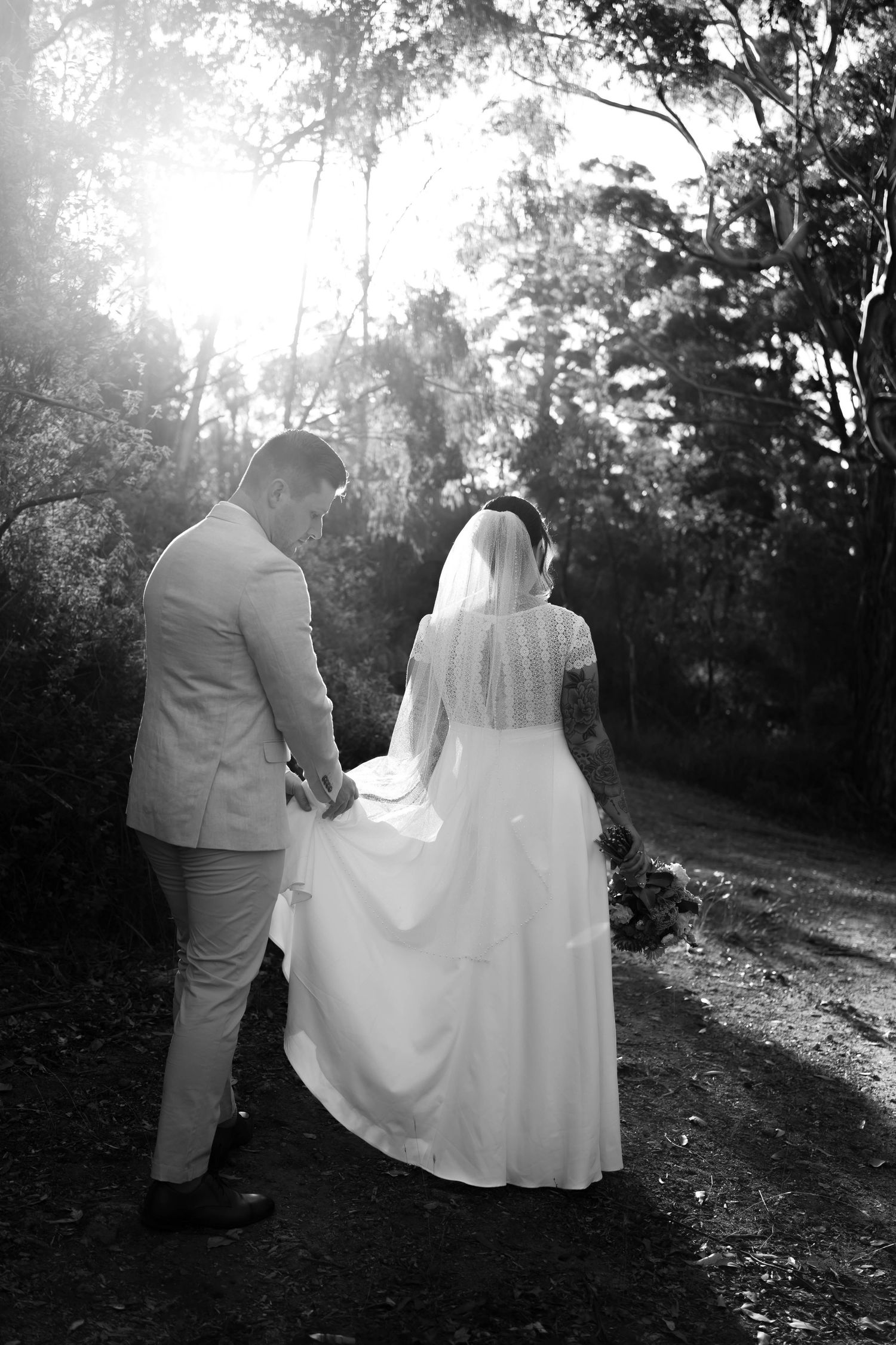 Black and white photo of bride and groom walking hand in hand through a sunlit forest path on their wedding day.