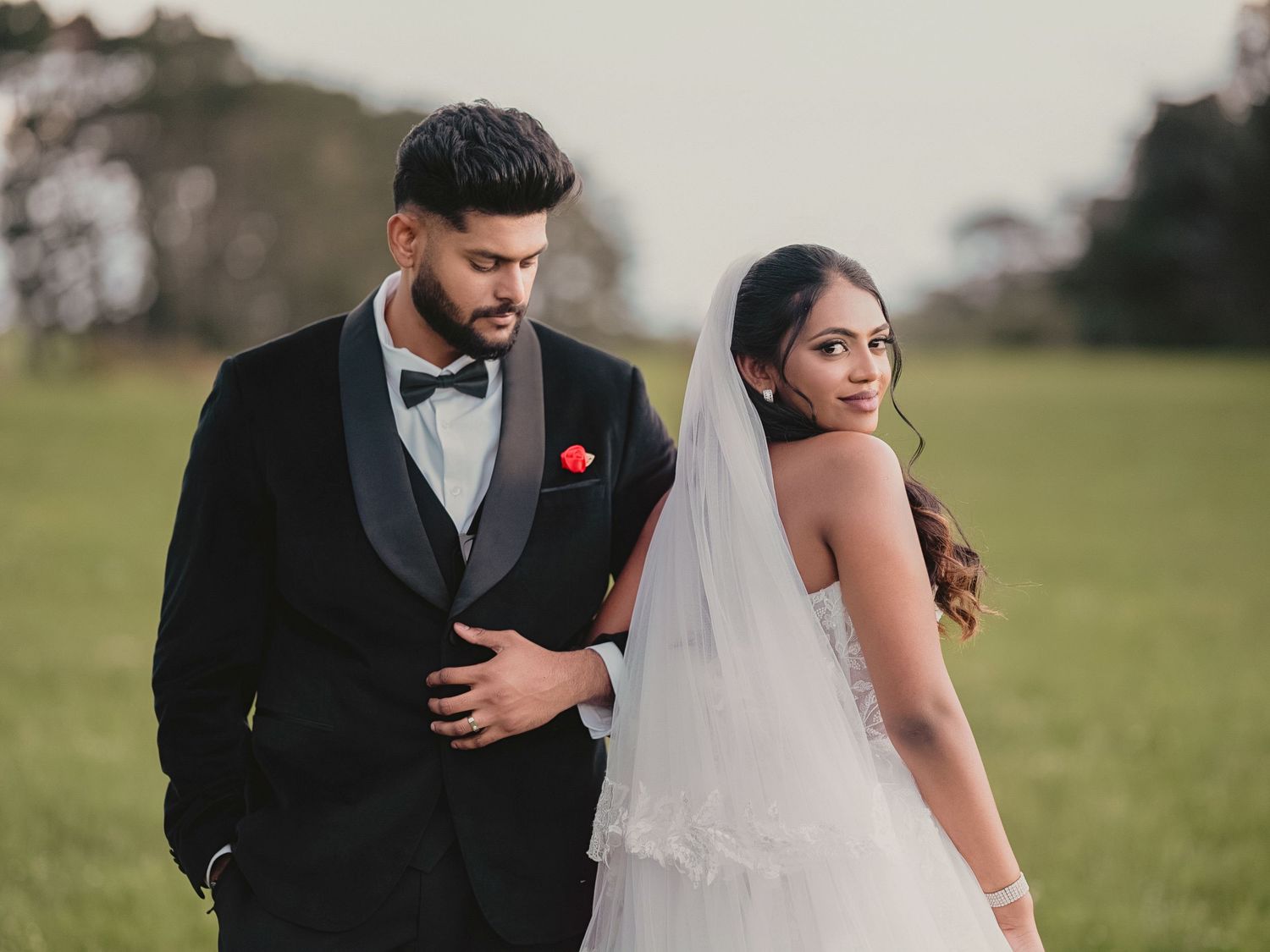 Bride in white gown and veil poses with groom in black tuxedo on lush green field at dusk.