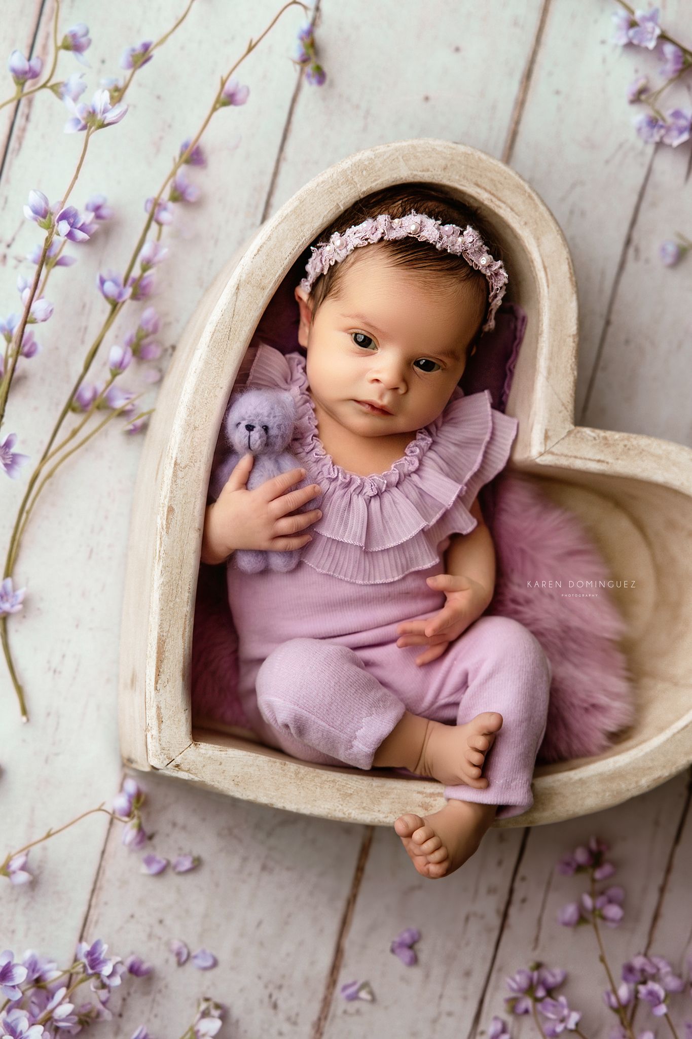 Newborn baby in purple outfit resting in heart-shaped wooden bowl with lavender flowers and floral headband.