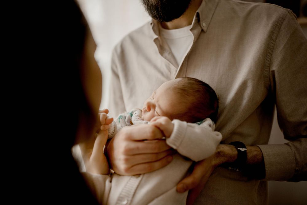 Un moment tendre d'un parent en chemise blanche berçant un nouveau-né endormi dans une douce lumière naturelle.