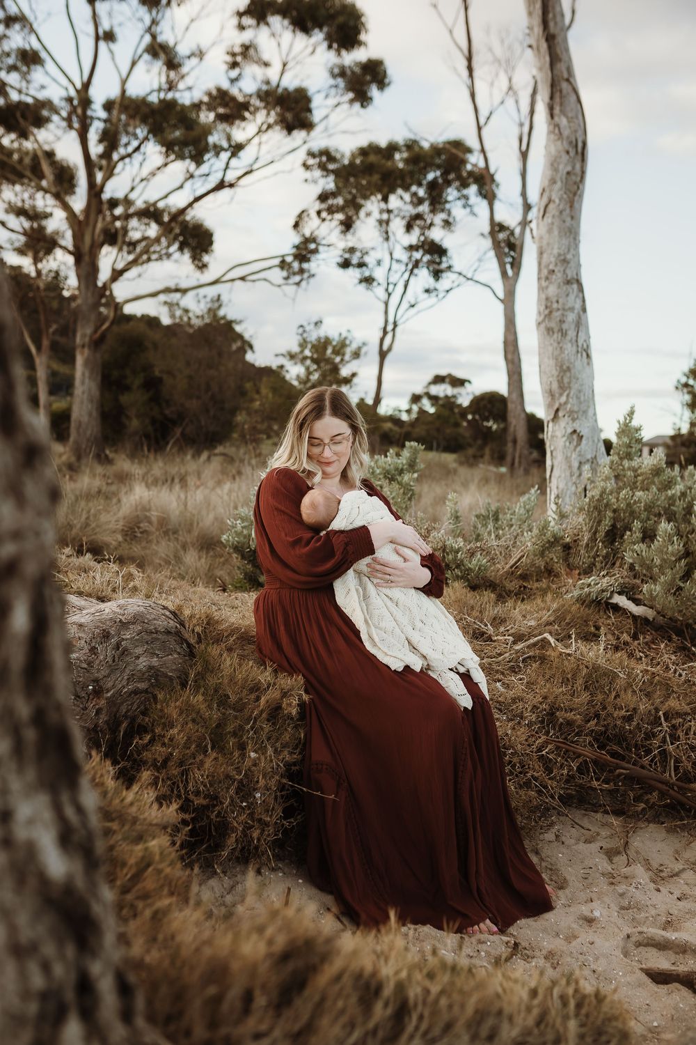 A mother in a dark red dress sits outdoors cradling her newborn baby, surrounded by native trees and golden grass.
