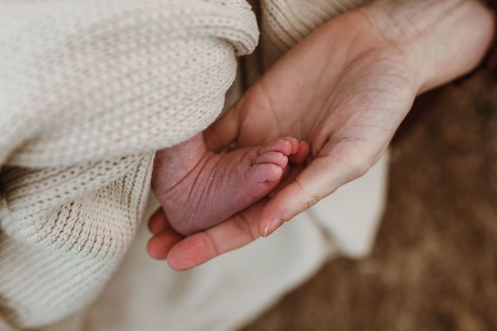 An adult hand gently holding a newborn baby's tiny foot wrapped in a soft cream knit blanket.