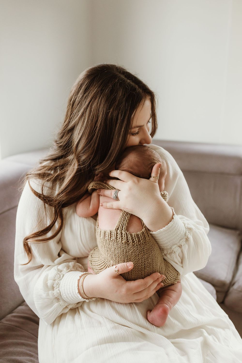 A mother with long brown hair tenderly holds her sleeping newborn baby on a couch.