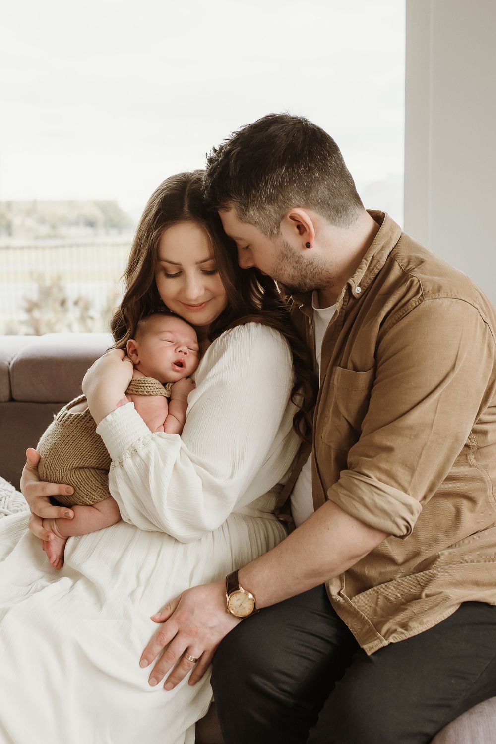 A couple tenderly holds their sleeping newborn baby, the mother in white and father in tan, sharing a loving family moment.