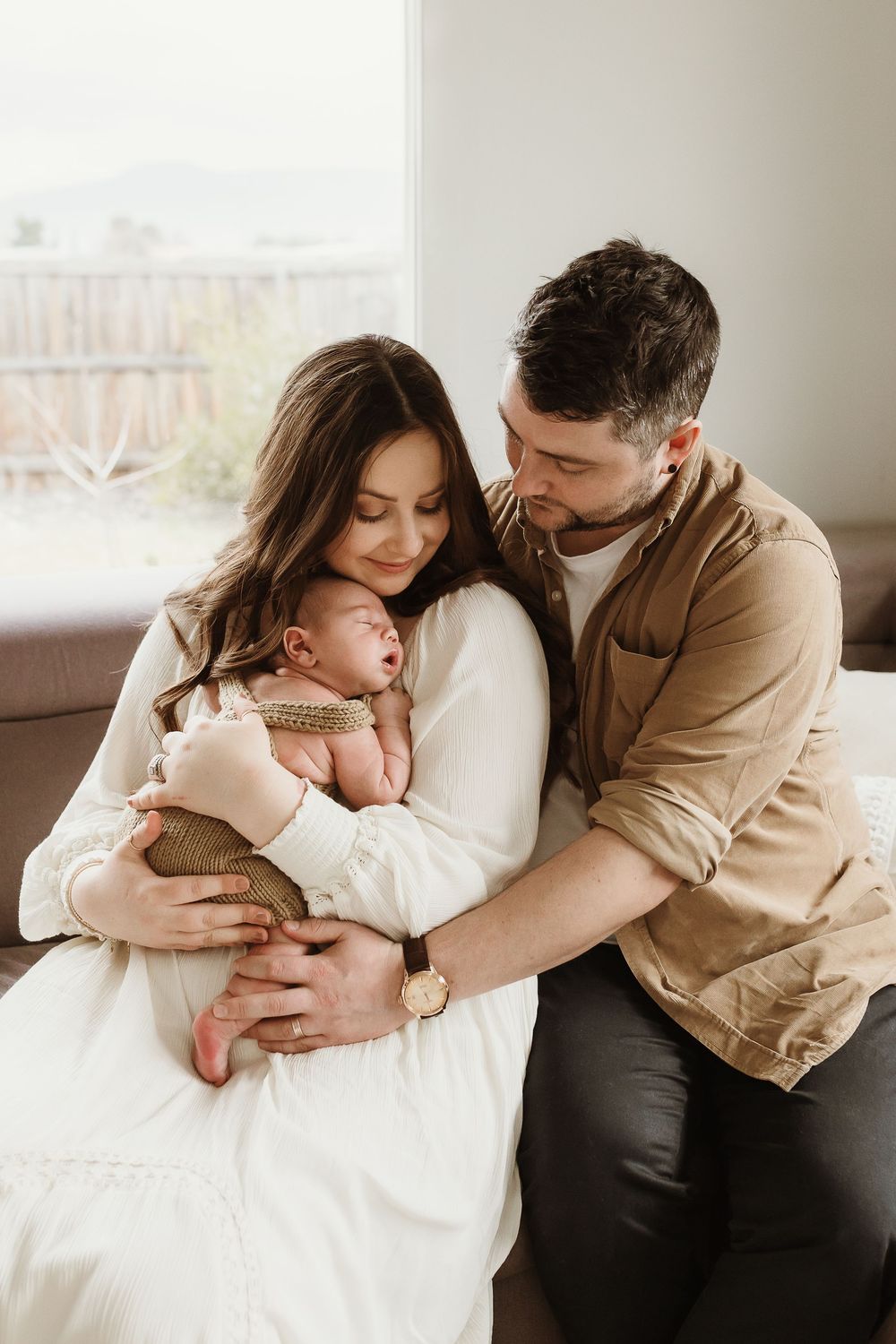 New parents tenderly cradling their newborn baby together on a white bed in a bright, cozy home setting.