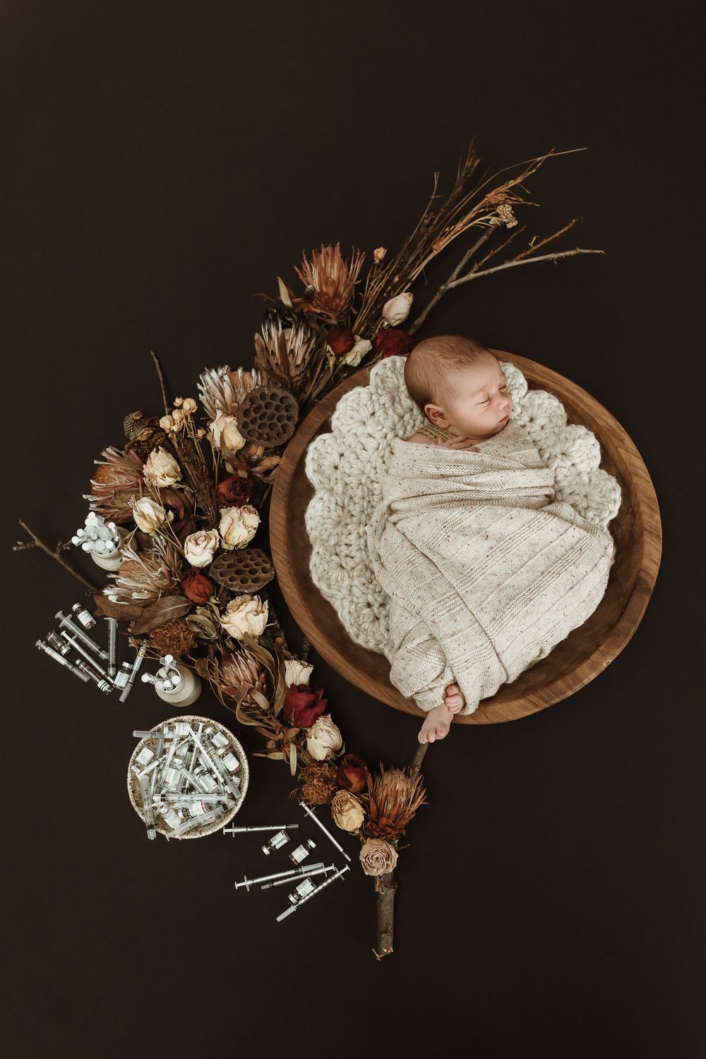 Sleeping newborn baby swaddled in cream knit wrap in a wooden bowl with dried floral arrangement.