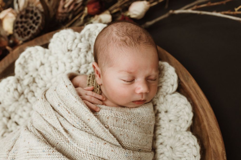 Sleeping newborn baby swaddled in cream knit wrap, nestled in wooden bowl with crochet blanket and dried floral backdrop.