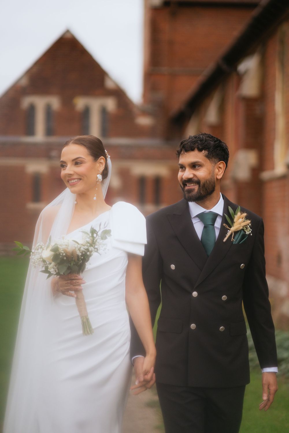 A wedding couple walks together outdoors in front of brick buildings, with the bride in white and groom in black suit.