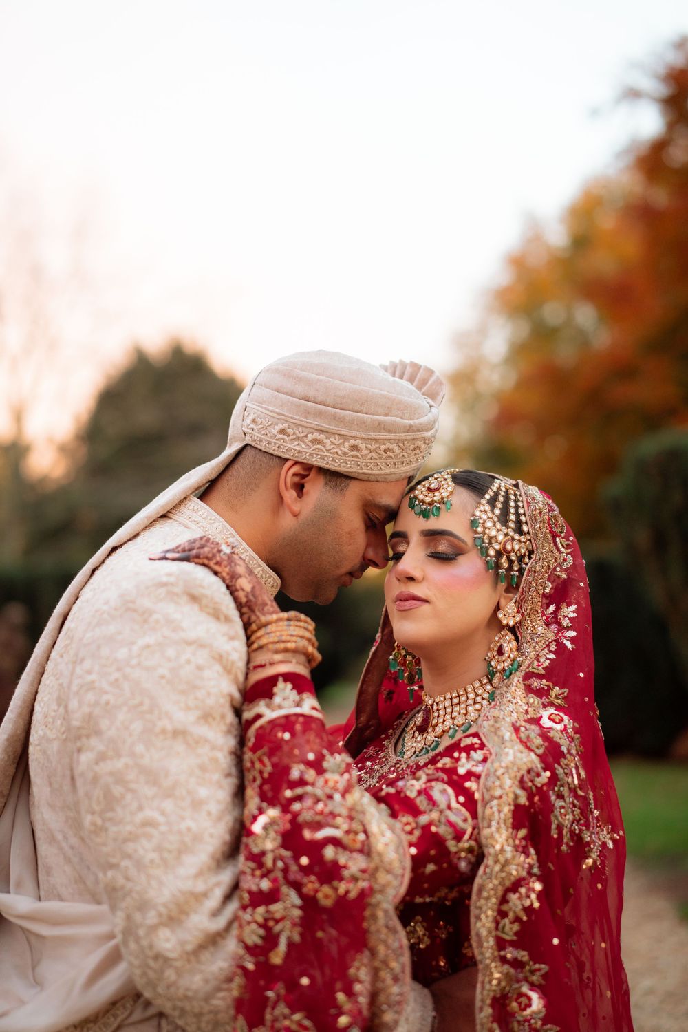A couple in traditional South Asian wedding attire embraces during an outdoor autumn photoshoot.