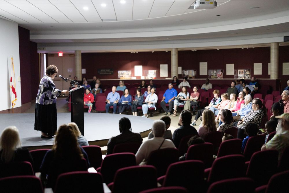 Woman speaking at podium to a seated audience in a red-chaired auditorium during a community event.