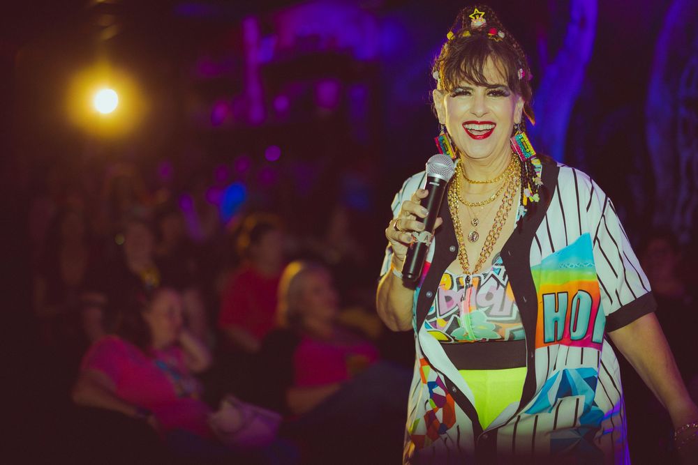 Performer in colorful jersey and gold jewelry sings into microphone on a lit stage.