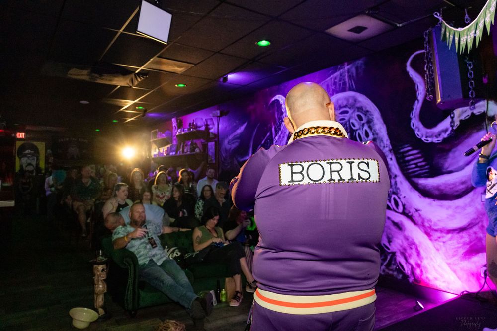 Performer in purple Boris jacket entertains crowd at dimly lit bar with octopus mural.