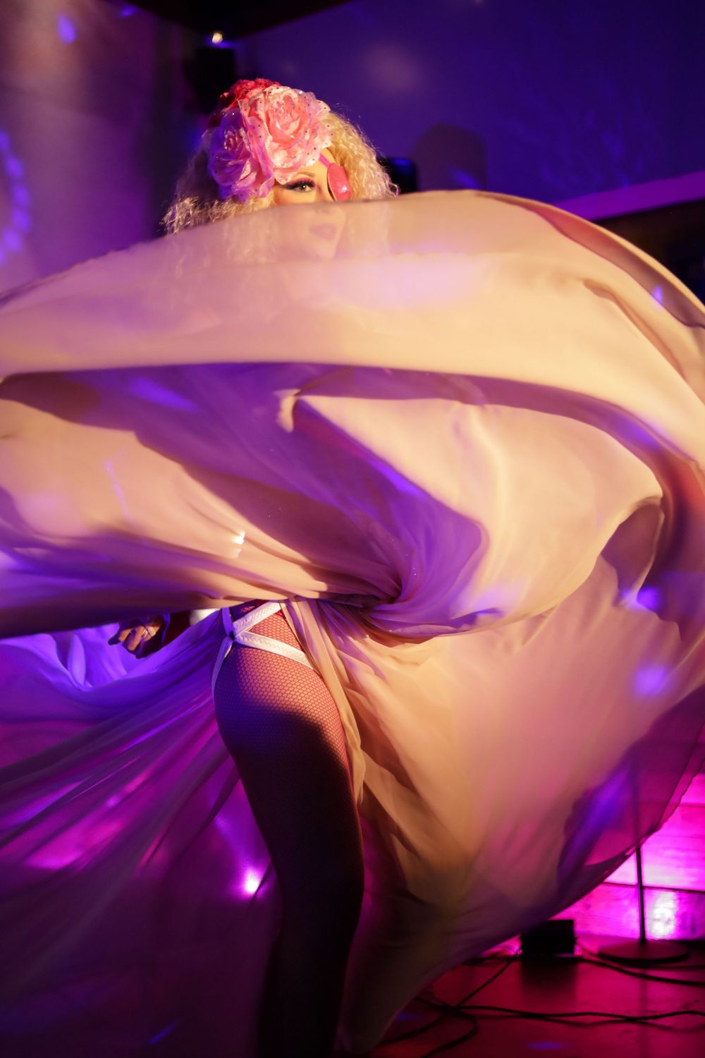 Blonde performer twirling flowing white fabric under purple stage lights in a dramatic dance.