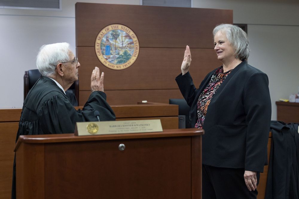 A judge administers an oath to a woman raising her right hand in a formal courtroom setting.