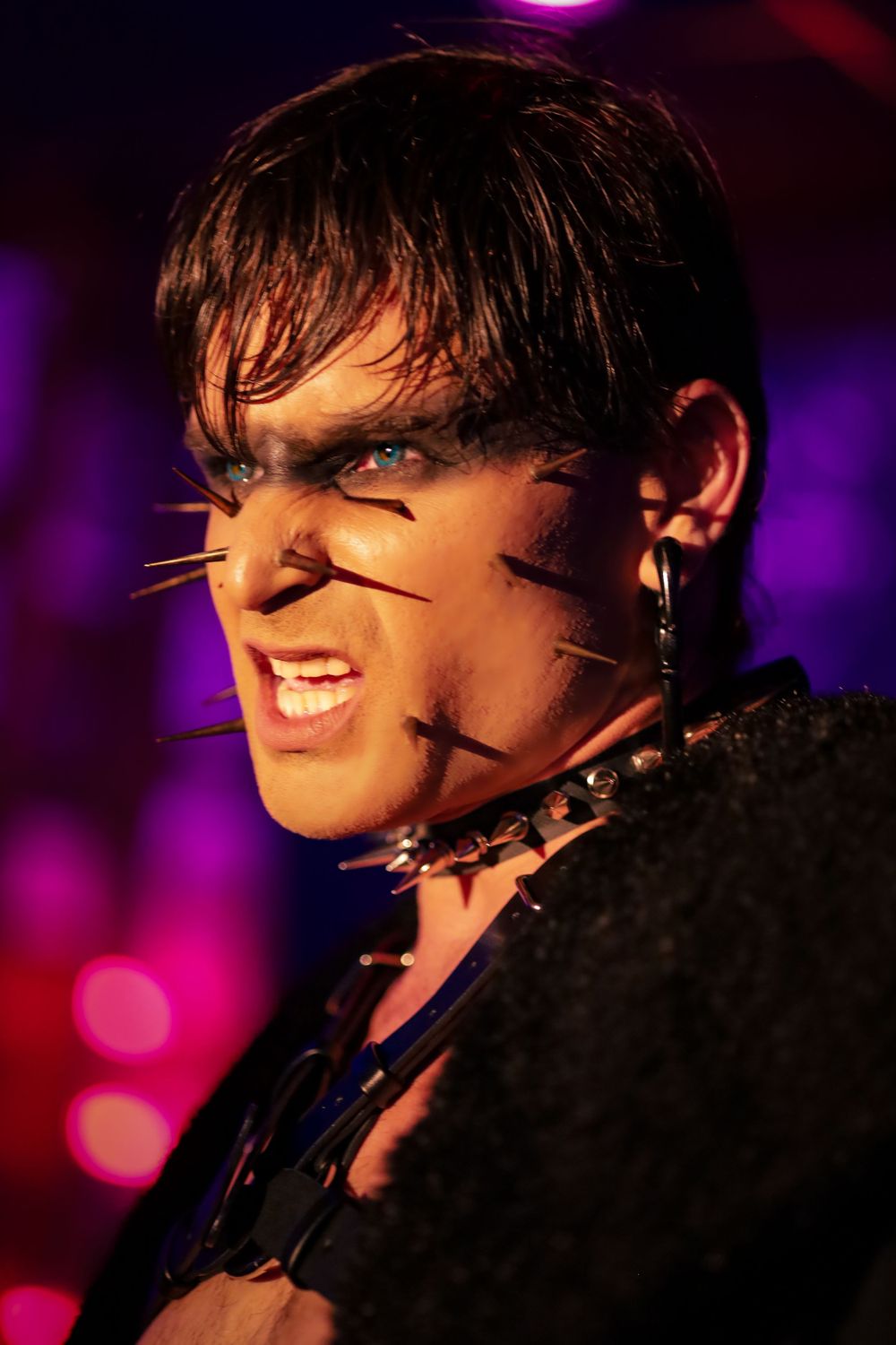 Performer with cat face paint, whiskers, and blue eyes snarls under purple stage lighting.