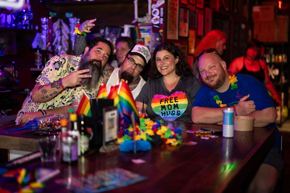 Group of friends smiling at a bar during Pride, wearing rainbow accessories and a Free Mom Hugs shirt.