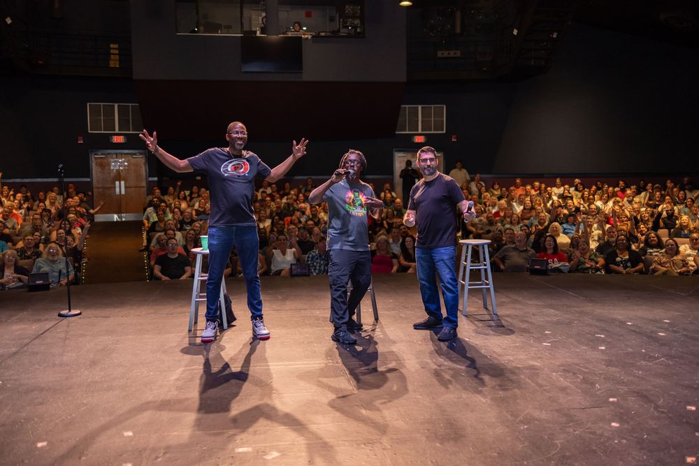 Three performers take a bow on stage before a large, enthusiastic crowd at an indoor comedy or variety show event.