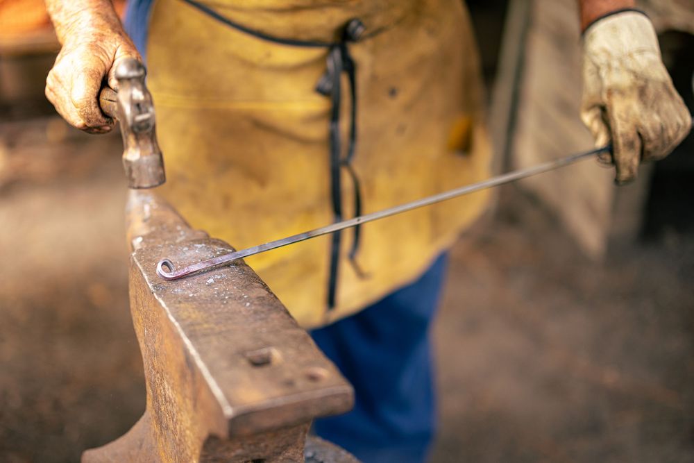 Blacksmith hammering metal rod on anvil while wearing protective gloves and yellow leather apron.