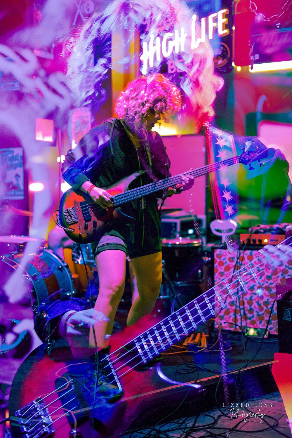 Bassist with curly pink hair performs under vibrant neon lights at a live music venue with High Life sign.