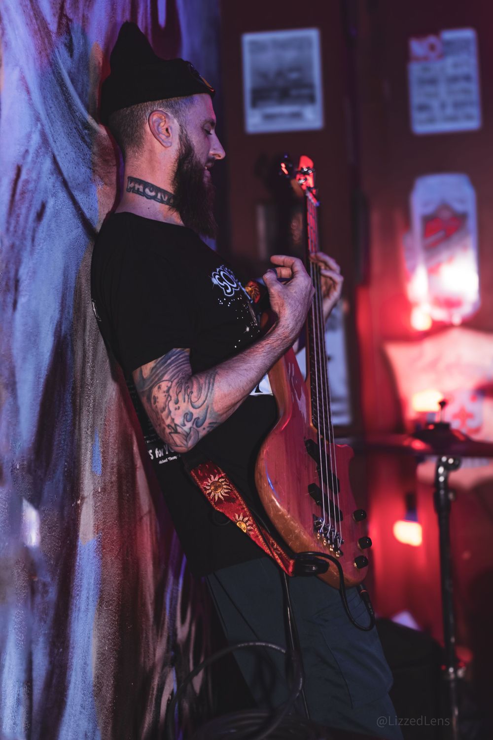 A tattooed musician plays an electric cello against a graffiti wall under moody red and purple stage lighting.