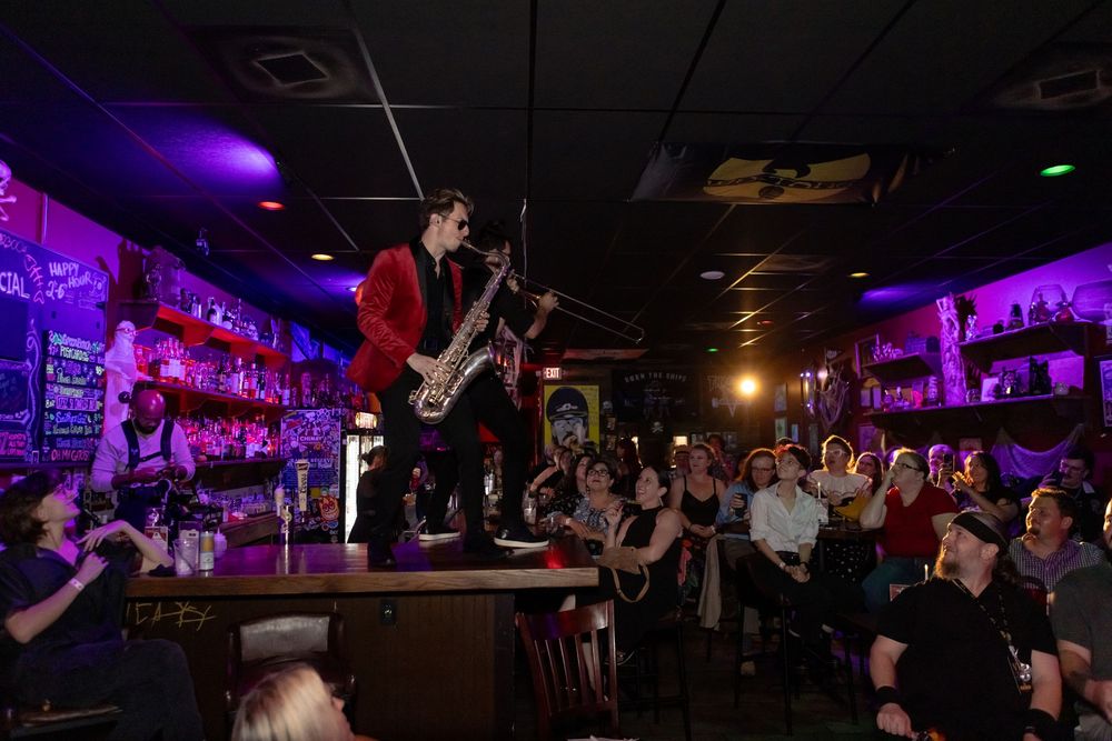 Saxophonist performs on a bar counter for a packed crowd in a purple-lit music venue.