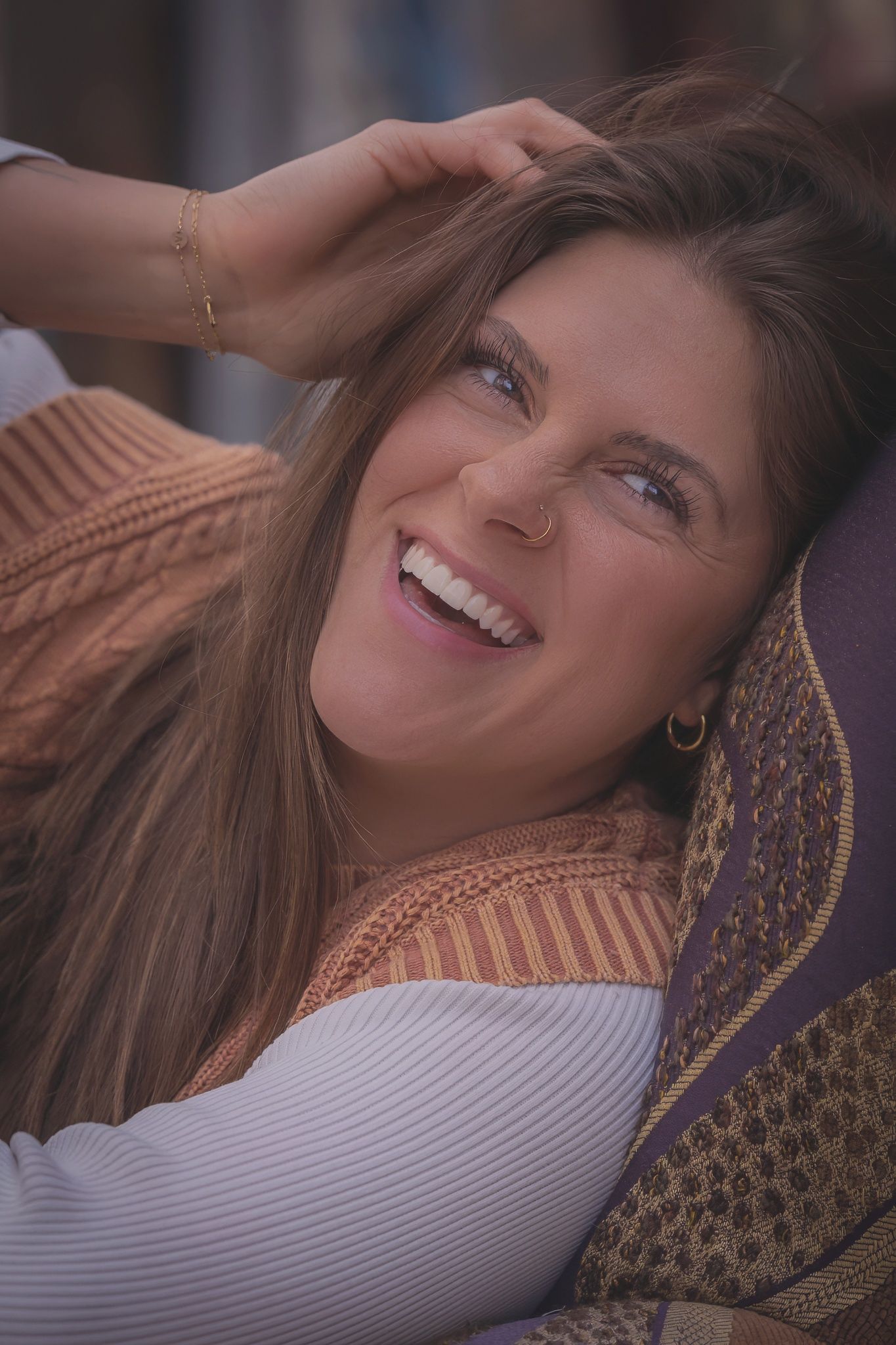 A joyful person with long brown hair wearing a tan sweater reclines while laughing against purple cushions.