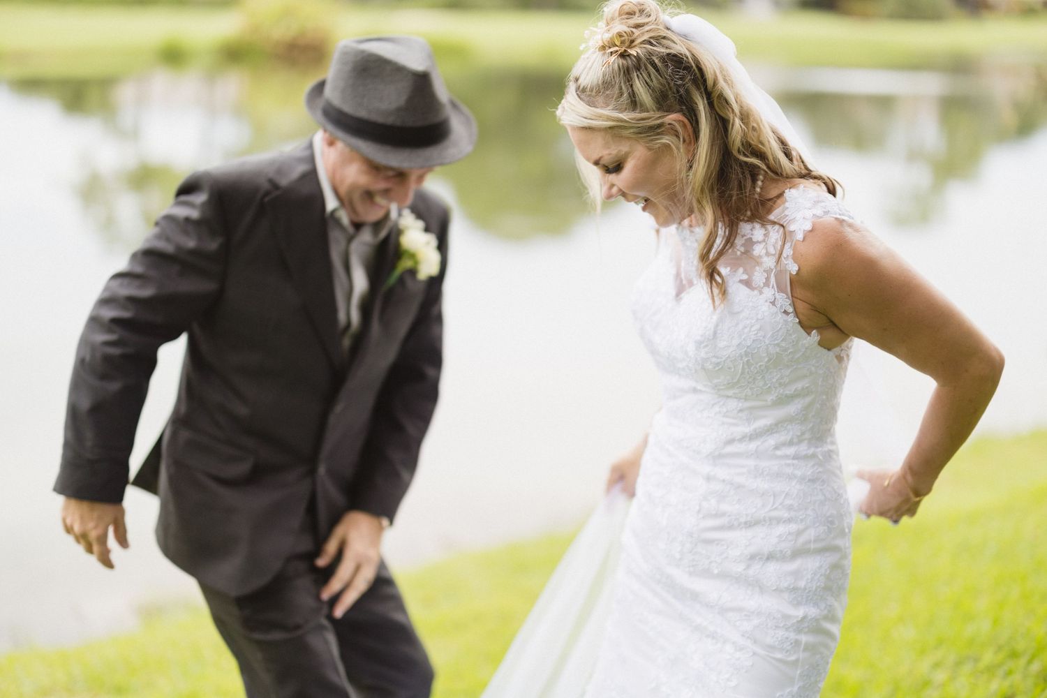 A playful wedding moment captured on grass near a pond with spontaneous dancing.