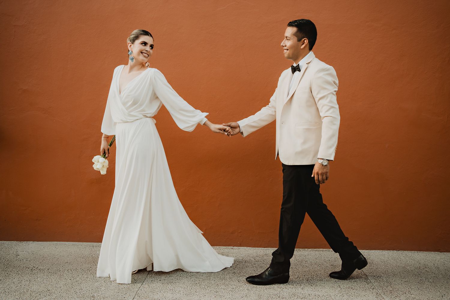 Pareja caminando tomada de la mano frente a un muro terracota durante una sesión de fotografía de pareja en Mazatlán, México