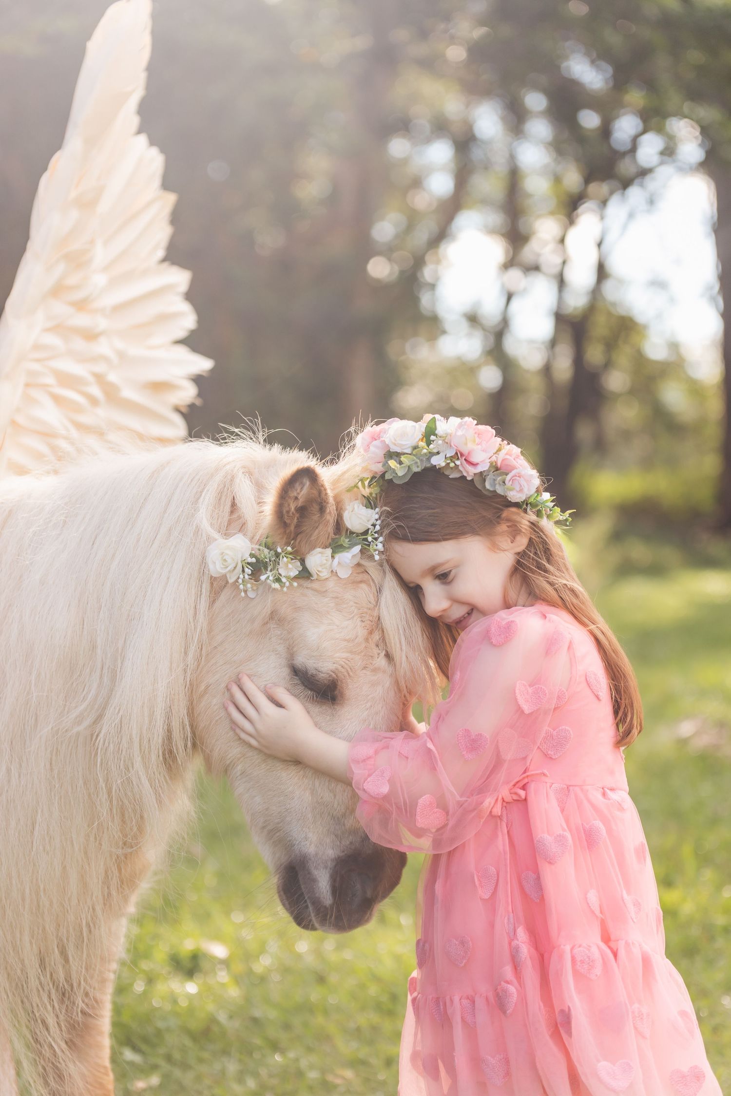 Child in pink dress shares tender moment with white horse in sunlit field.