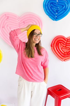 Person in pink sweater and yellow beanie poses against wall with colorful heart decorations.