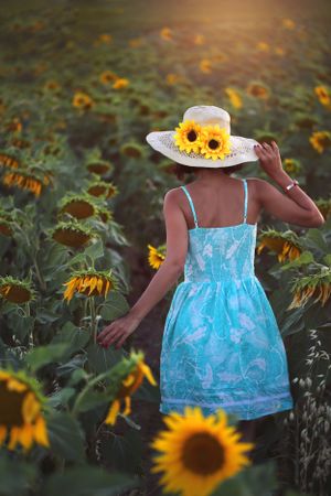 Artistic back view of someone in a blue dress and sunflower-adorned hat walking through a sunflower field.
