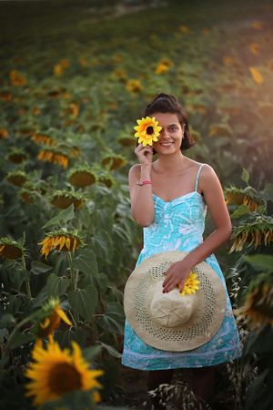 Cheerful summer scene with someone in a blue dress holding a straw hat among blooming sunflowers.
